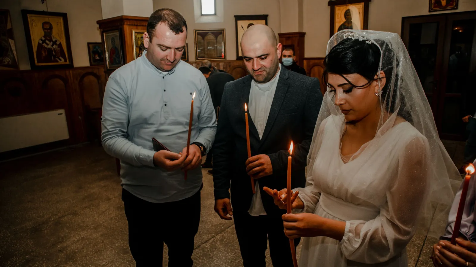 A veiled bride and two men holding lit wax candles during an Orthodox church wedding, with icons on the walls